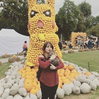 Katie Powell photograph. A brown-haired woman holds a cat while standing in front of a Pikachu made of gourds. 
